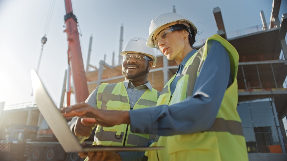 Two construction workers wearing hard hats and safety vests review plans on a laptop at a building site, highlighting the importance of engineering inspections NYC, with a crane and partially constructed building in the background on a sunny day.