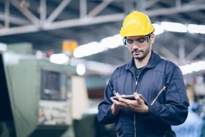 A male worker wearing a yellow hard hat, safety glasses, and a blue jumpsuit stands in a factory, using a digital tablet to conduct engineering inspections NYC, with machinery visible in the background.