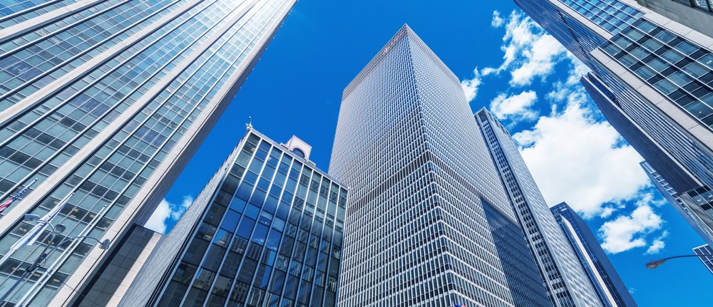 Tall modern skyscrapers with glass windows surround the viewer, reaching toward a bright blue sky with scattered white clouds, setting the scene for professional engineering inspections NYC from a low-angle perspective.