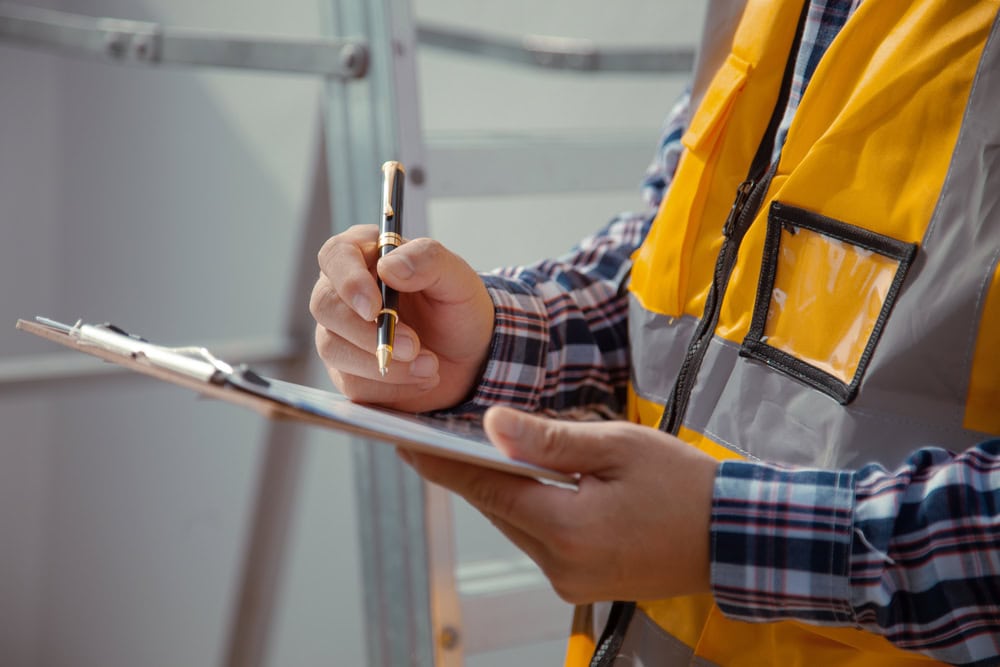 A person in a yellow safety vest and plaid shirt writes on a clipboard, standing near a metal ladder—capturing the diligence of engineering inspections NYC. Only their torso and hands are visible.