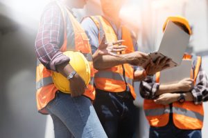 Three construction workers in orange safety vests discuss engineering inspections NYC outdoors. One holds a yellow hard hat, another points at a clipboard, all under bright sunlight. Their faces are not visible.