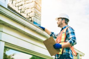 A construction worker in a hard hat, safety vest, and gloves holds a clipboard while performing engineering inspections NYC on the roof and gutter of a house on a sunny day.