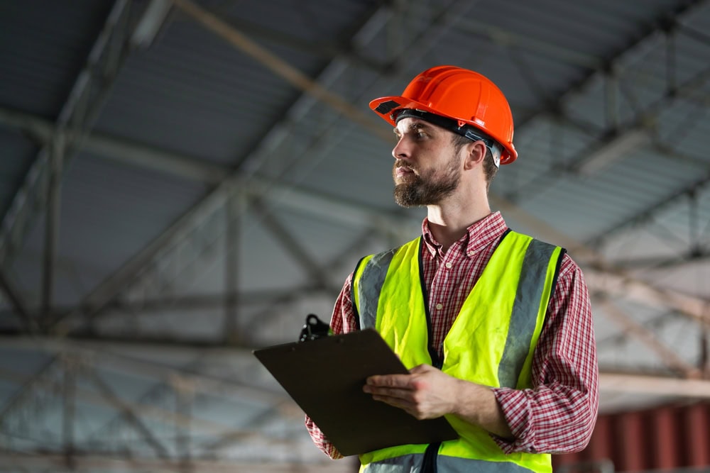 A construction worker in a reflective safety vest and orange hard hat holds a clipboard and looks to the side inside a large industrial building, highlighting the importance of engineering inspections NYC.