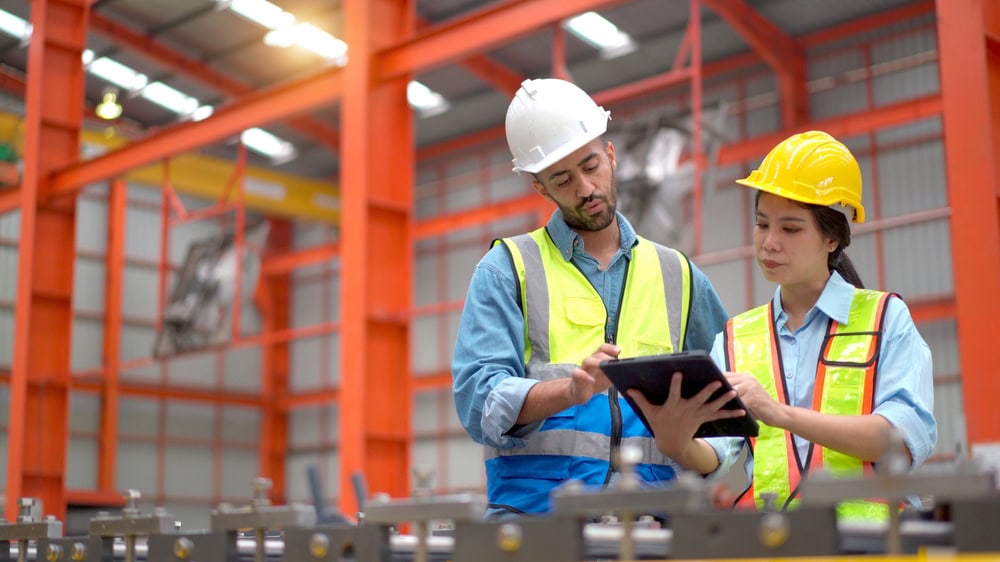 Two workers in safety vests and helmets stand in a factory, reviewing information on a tablet together—demonstrating the precision required for engineering inspections NYC. Orange industrial structures and equipment fill the background.