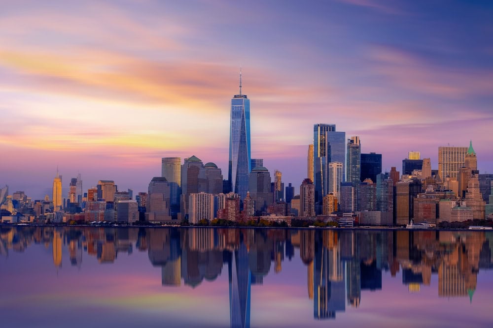 The New York City skyline at sunset, featuring tall skyscrapers including One World Trade Center, is beautifully reflected on calm water—an inspiring scene for engineering inspections NYC professionals ensuring the city’s safety and brilliance.