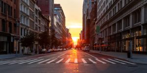 A city street at sunset with sunlight shining between tall buildings, reflecting on the road. Cars are parked along both sides, and the mostly empty street hints at recent engineering inspections NYC ensuring safety and compliance.