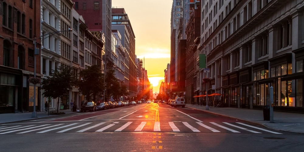 A city street at sunset with sunlight shining between tall buildings, reflecting on the road. Cars are parked along both sides, and the mostly empty street hints at recent engineering inspections NYC ensuring safety and compliance.