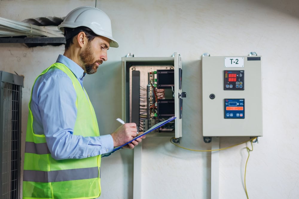 A man in a safety vest and hard hat writes on a clipboard while inspecting electrical control panels mounted on a wall during engineering inspections NYC.