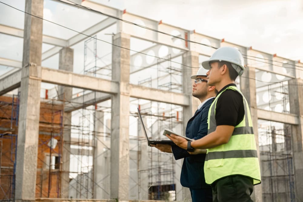 Two construction workers wearing safety helmets and vests stand outdoors at a building site, holding a laptop and a tablet as they conduct engineering inspections NYC, observing the steel frame structure under construction in the background.