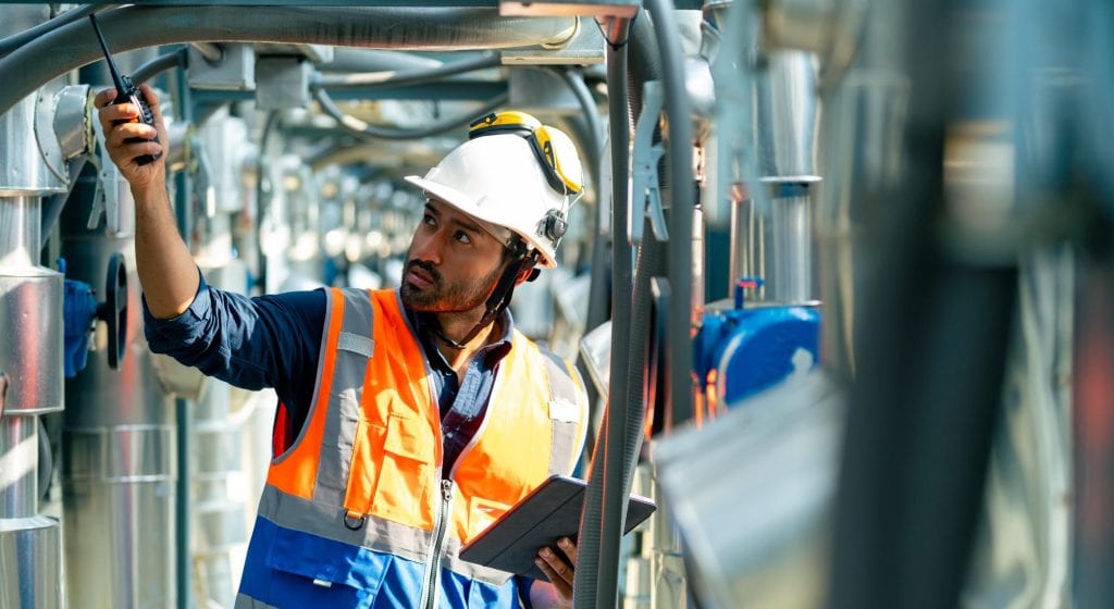 A worker in a hard hat, safety goggles, and orange vest uses a walkie-talkie and holds a tablet while performing engineering inspections NYC at an industrial facility.