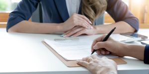 A person is signing a contract for inspection services NYC on a clipboard at a desk, while another person with folded hands sits across from them. A calculator and smartphone are also visible on the desk.