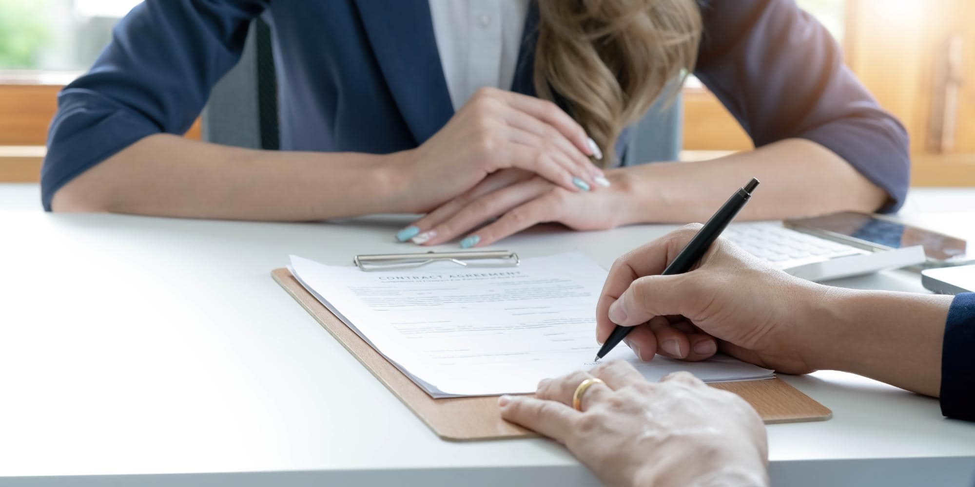 A person is signing a contract for inspection services NYC on a clipboard at a desk, while another person with folded hands sits across from them. A calculator and smartphone are also visible on the desk.