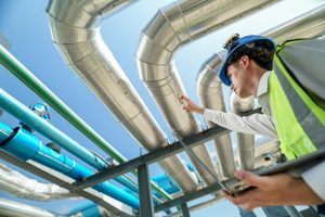 A worker in a hard hat and safety vest inspects large metal pipes outdoors under a blue sky, holding a tablet and reaching up toward the pipes—demonstrating expert engineering inspections NYC relies on.