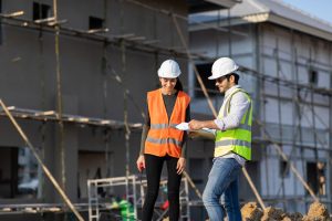 Two construction workers in safety vests and hard hats discuss project plans outside a building under construction. Scaffolding is visible in the background, highlighting the importance of engineering inspections NYC for safety and compliance.