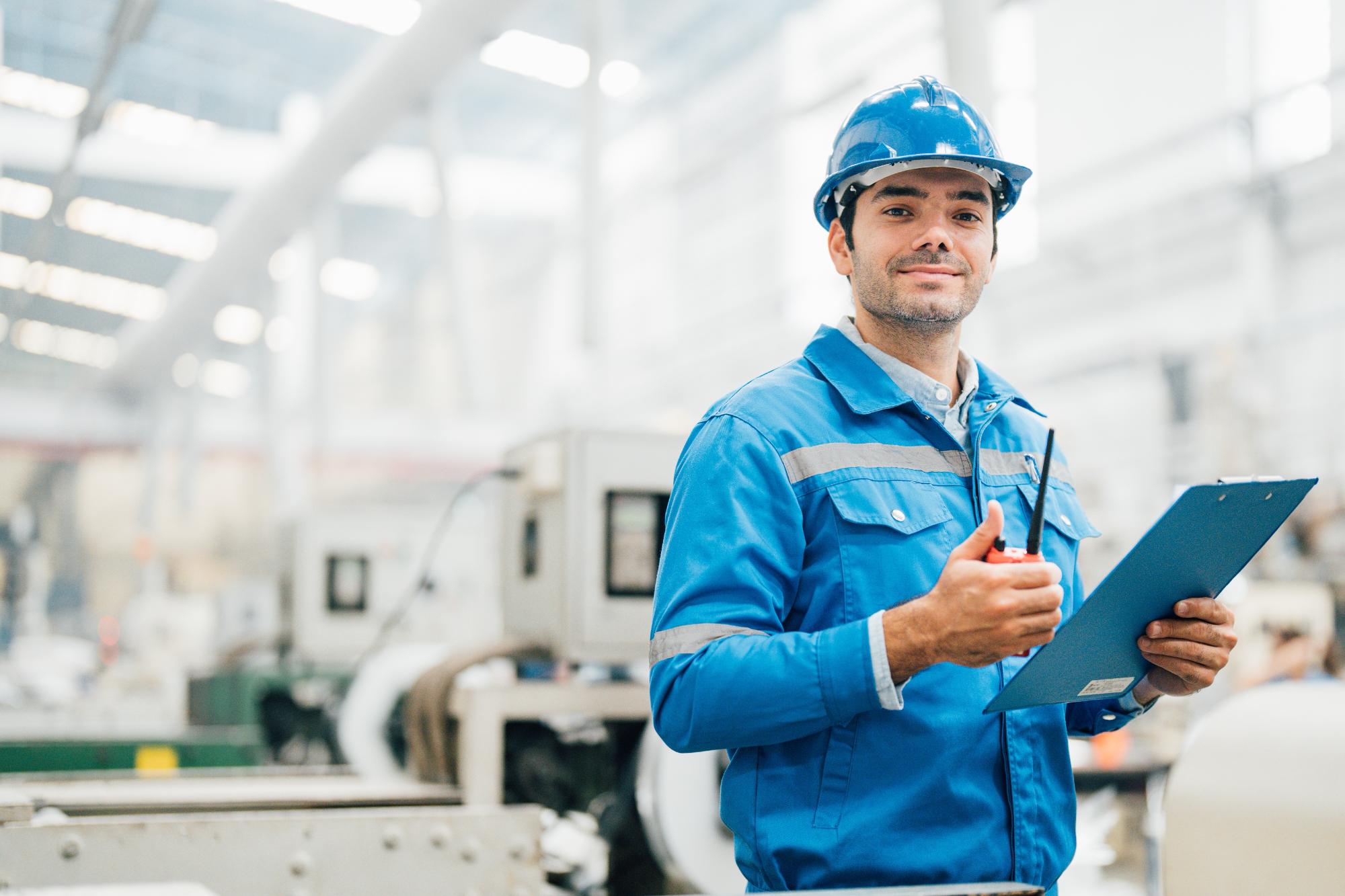 A factory worker in a blue uniform and helmet holds a clipboard and walkie-talkie, smiling as he conducts engineering inspections NYC in a brightly lit industrial setting with machines in the background.