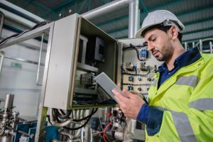A male engineer in a high-visibility jacket and helmet uses a tablet while performing engineering inspections NYC on electrical equipment in an industrial facility.