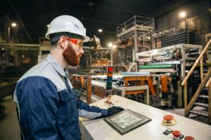 A man wearing a hard hat and safety glasses operates a touchscreen control panel in an industrial factory setting, surrounded by machinery—a scene typical of engineering inspections NYC professionals conduct daily.