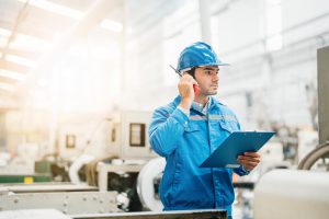 A male worker in a blue uniform and hard hat stands in a factory, holding a clipboard and speaking into a walkie-talkie, conducting engineering inspections NYC amid machinery and industrial equipment.