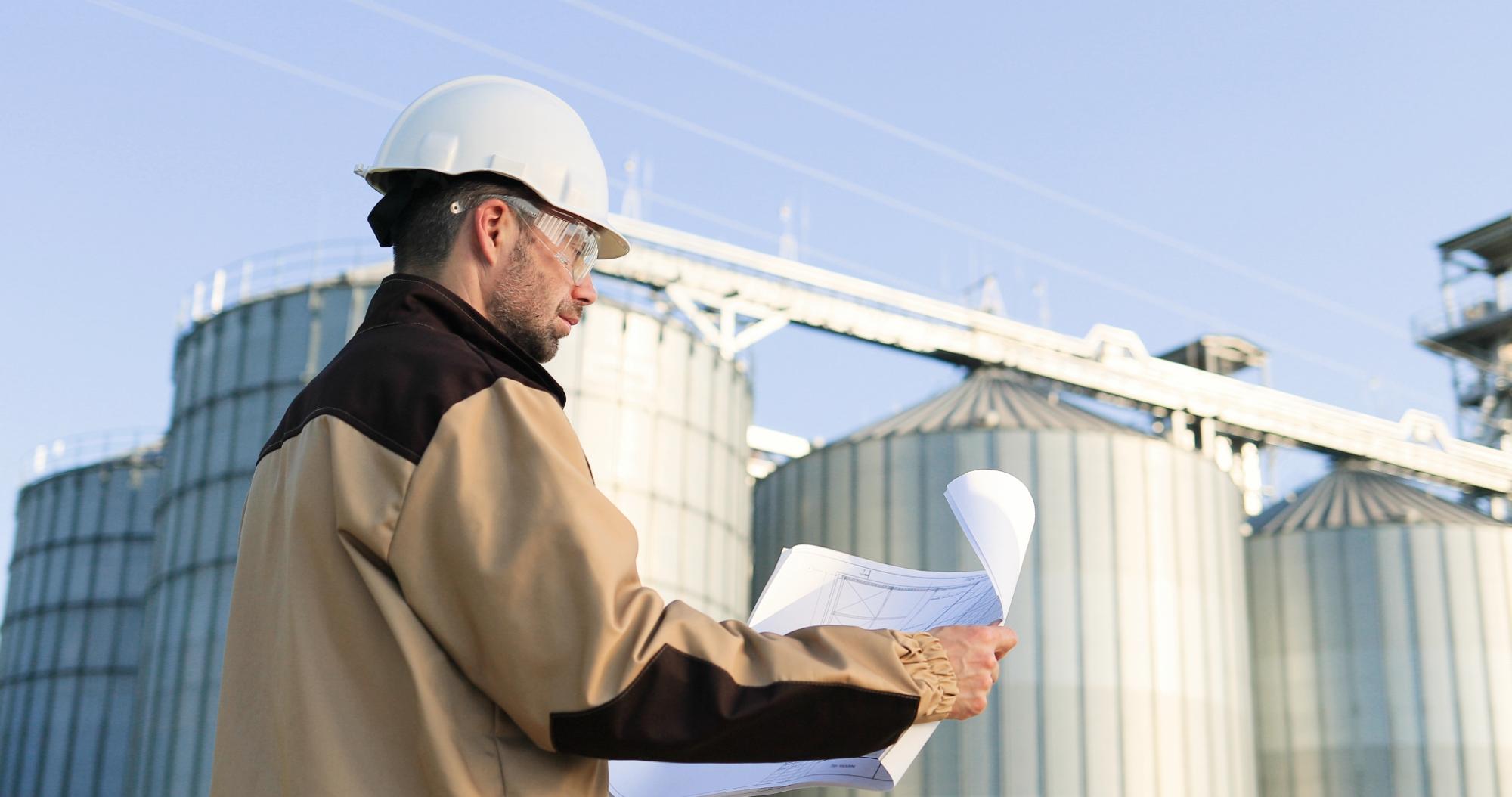 A person wearing a hard hat and safety glasses examines blueprints outdoors in front of large industrial silos, representing the precision required for engineering inspections NYC under a clear blue sky.