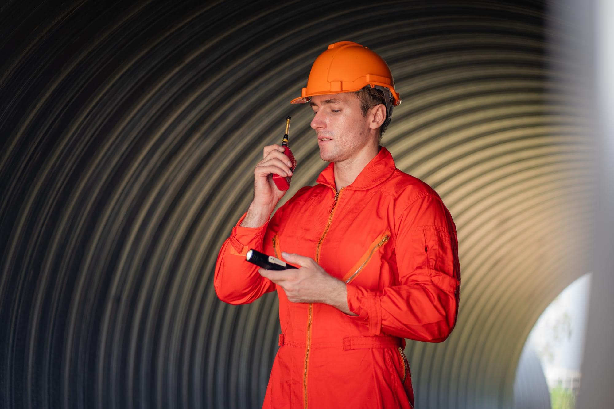 A construction worker in an orange jumpsuit and hard hat uses a walkie-talkie while standing inside a large, curved, metallic tunnel during engineering inspections NYC.