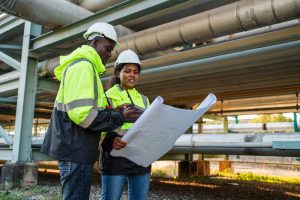 Two engineers in safety jackets and helmets review blueprints together at an industrial site with large metal pipes, highlighting the importance of engineering inspections NYC for safety and compliance.