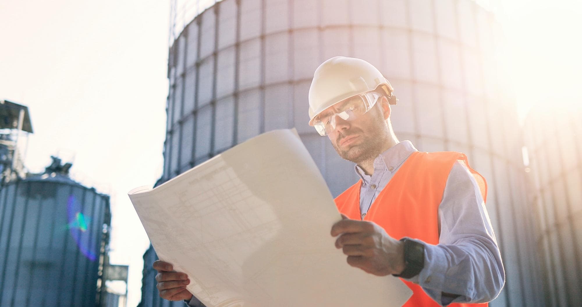A construction worker wearing a white hard hat, safety glasses, and an orange vest examines blueprints outdoors, highlighting the importance of engineering inspections NYC near large industrial silos under bright sunlight.