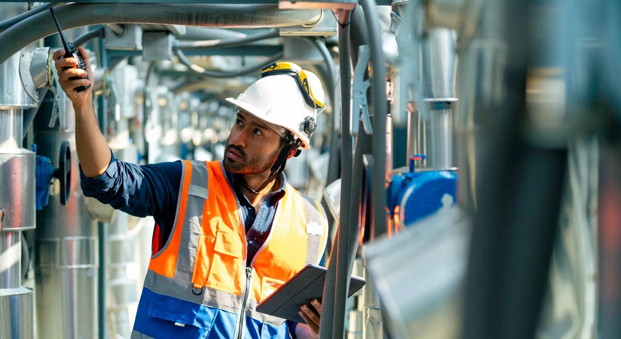 A worker in a white hard hat and orange safety vest conducts special inspections NYC, holding a walkie-talkie and tablet while closely examining industrial pipes surrounded by metallic equipment.