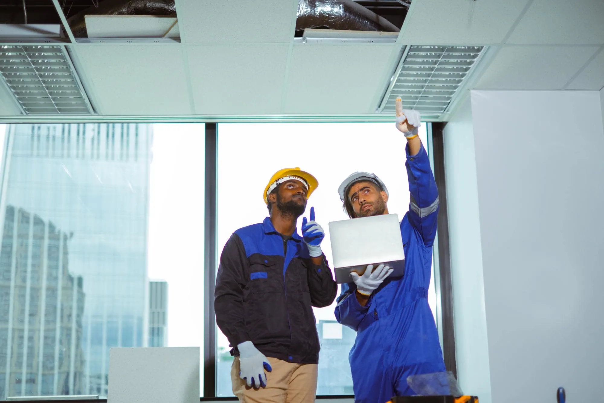 Two workers in safety gear stand in an office, looking up and pointing at a ceiling panel with exposed ducts, one holding a laptop—conducting special inspections NYC. Large windows reveal city buildings in the background.