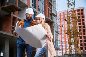Two construction workers wearing hard hats examine blueprints at a building site, with a crane and partially constructed building visible in the background during special inspections NYC projects.