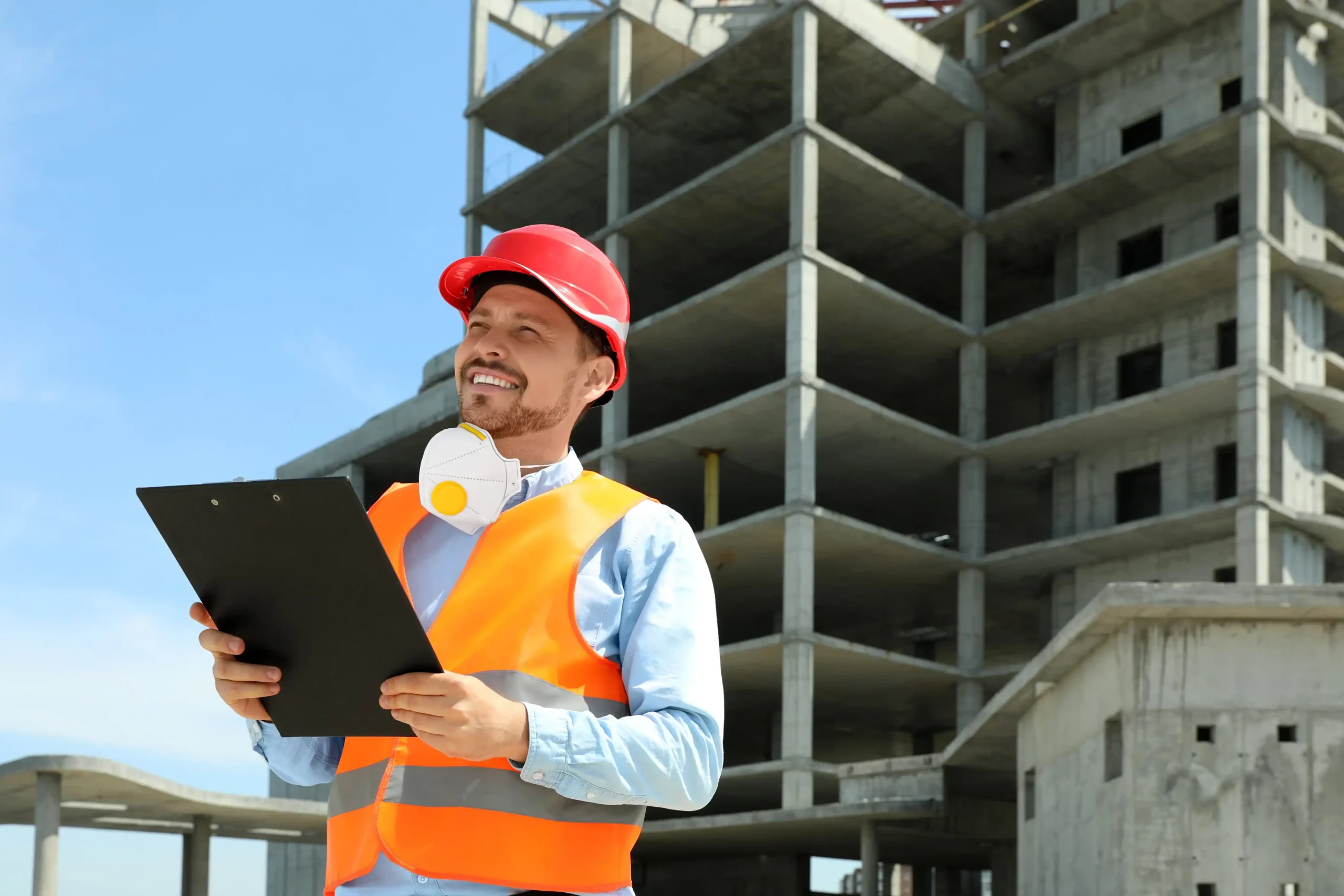 A man wearing a red hard hat, orange safety vest, and face mask around his neck holds a clipboard while smiling in front of a multi-story concrete building under construction for a Special Inspection Agency NY project.